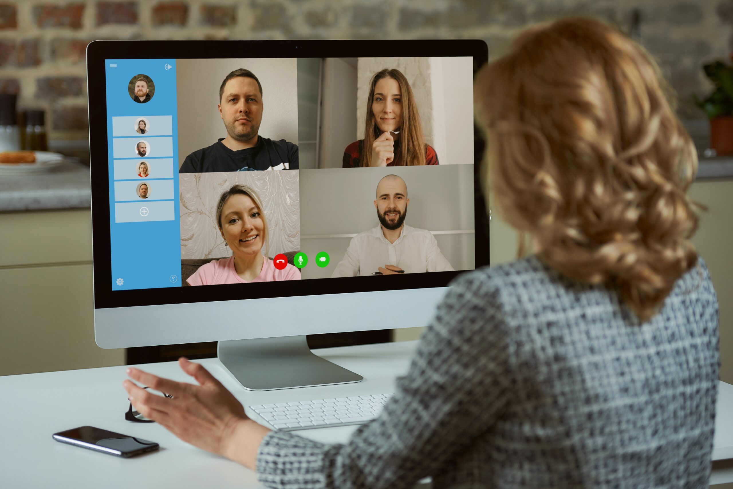 A back view of a woman who works remotely listening to reports of employees on an online briefing in her studio. A female teacher listening to students on a video call on a desktop computer at home.