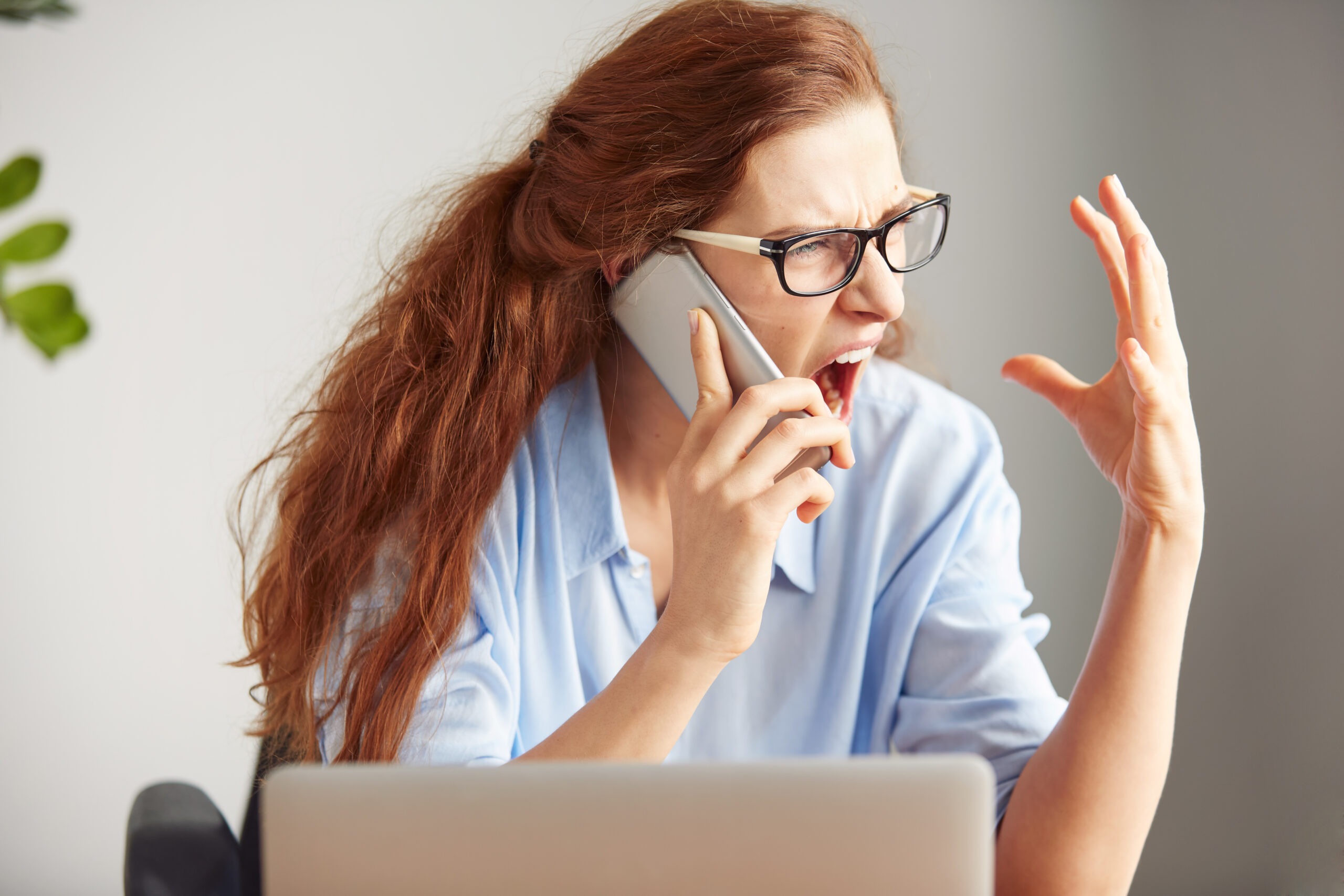 Headshot of a young female boss shouting with anger on the cell phone while sitting at the desk in her office. Portrait of an angry businesswoman screaming on the mobile phone. Negative human emotions, facial expressions