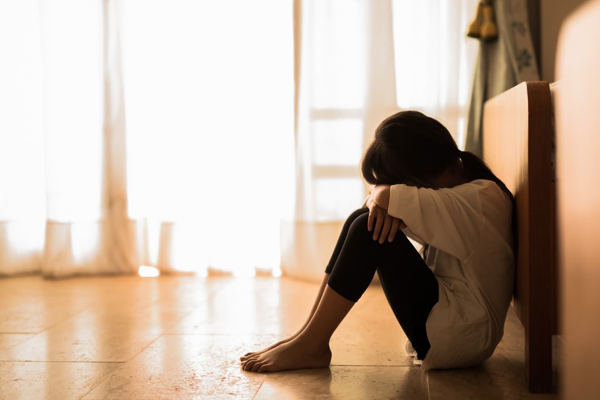 A victim of domestic violence sits crouched on the floor with her knees drawn up and her face in her arms.