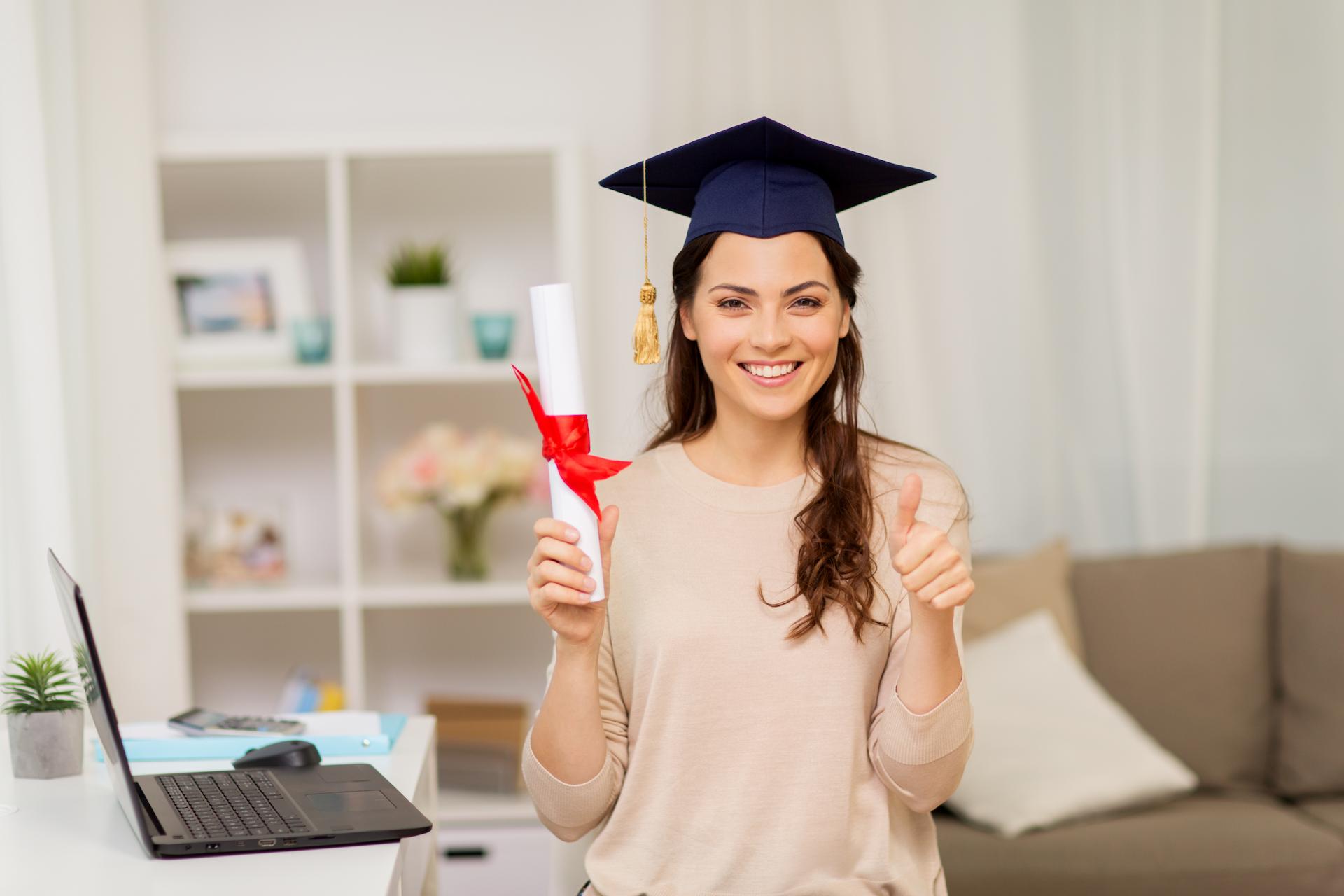 A smiling young woman at home wearing a graduation cap and holding her college degree gives a thumbs up.