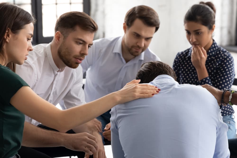An addiction counselor speaks with a troubled client as others in the group offer comfort.