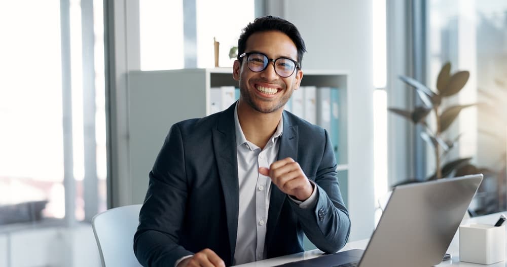 A smiling business administration sits at his desk working on his laptop.