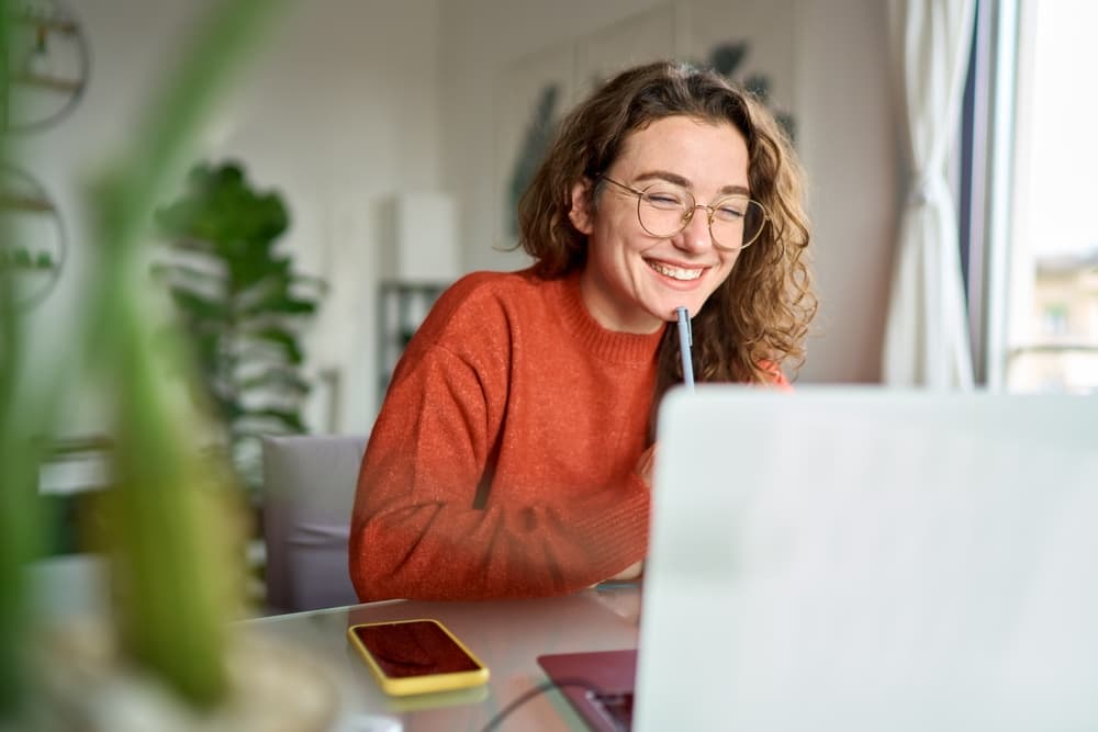 A smiling young woman takes notes while watching a lecture on her laptop.