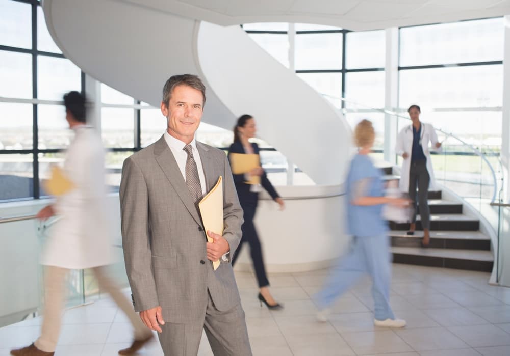 A hospital administrator smiles at the camera while standing in a busy hospital hallway.