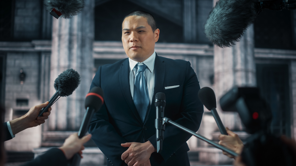 A man in a suit and tie stands addressing reporters holding microphones on the steps of a courthouse building.