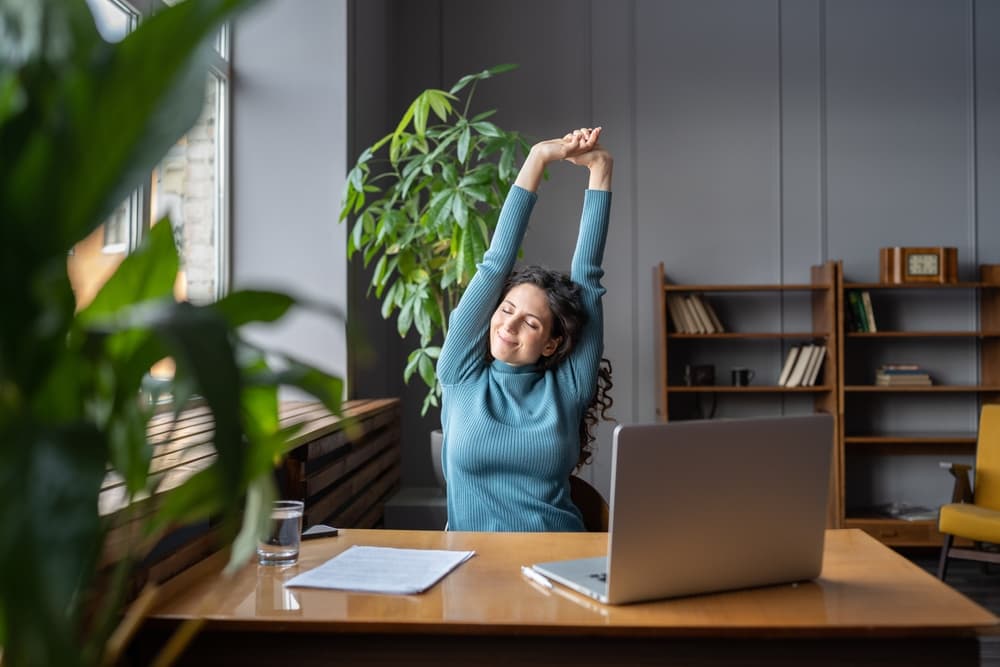 A happy, smiling worker stretches while taking a break at her desk.