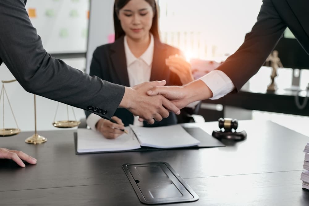 Men shake hands in the foreground as a businesswoman sits at her desk and writes in a notebook in the background.