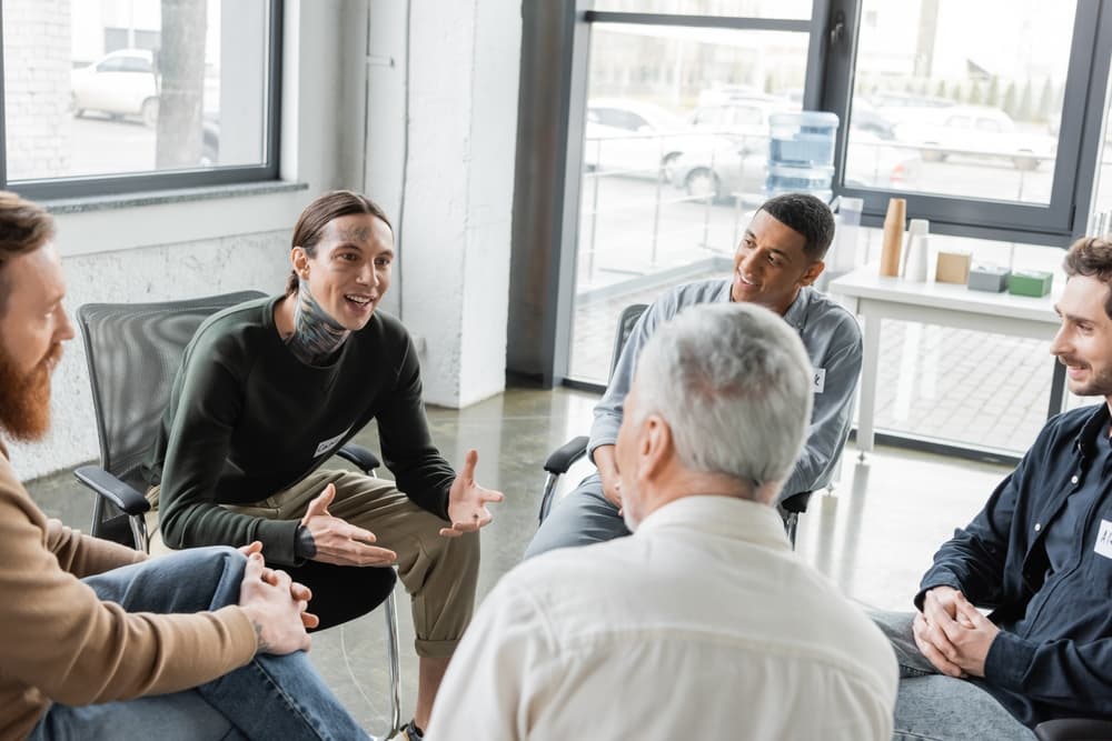 A smiling man with distinguishing neck tattoos shares with a small addiction counseling support group.