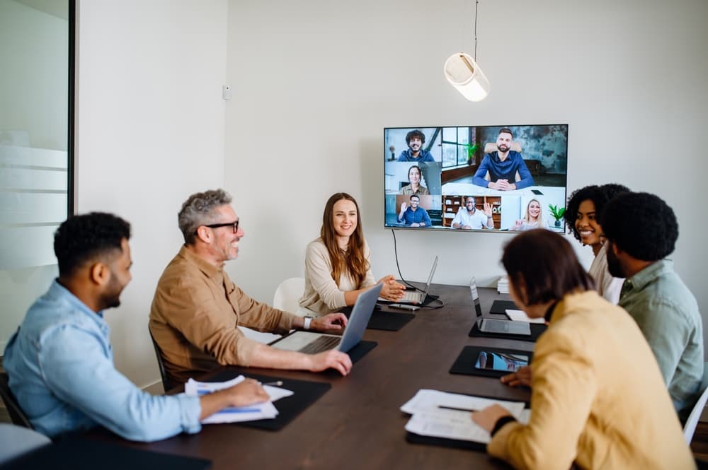A hybrid business team meets virtually and in an office meeting room.