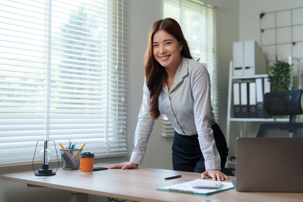 A smiling businesswoman stands at her office desk.