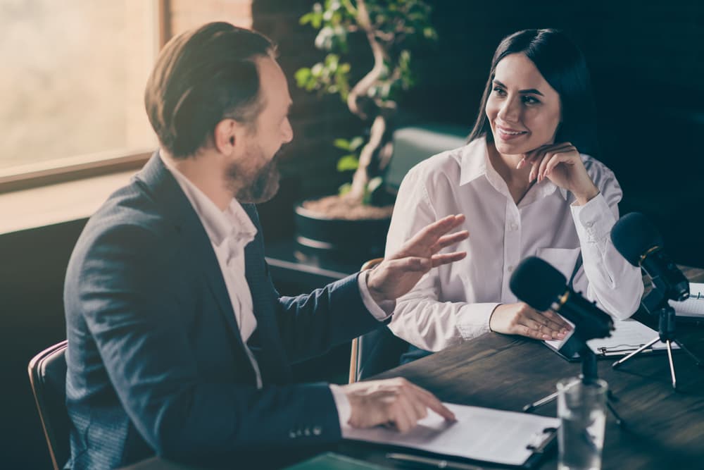 A male and a female speak with each other into microphones in a professional office setting.