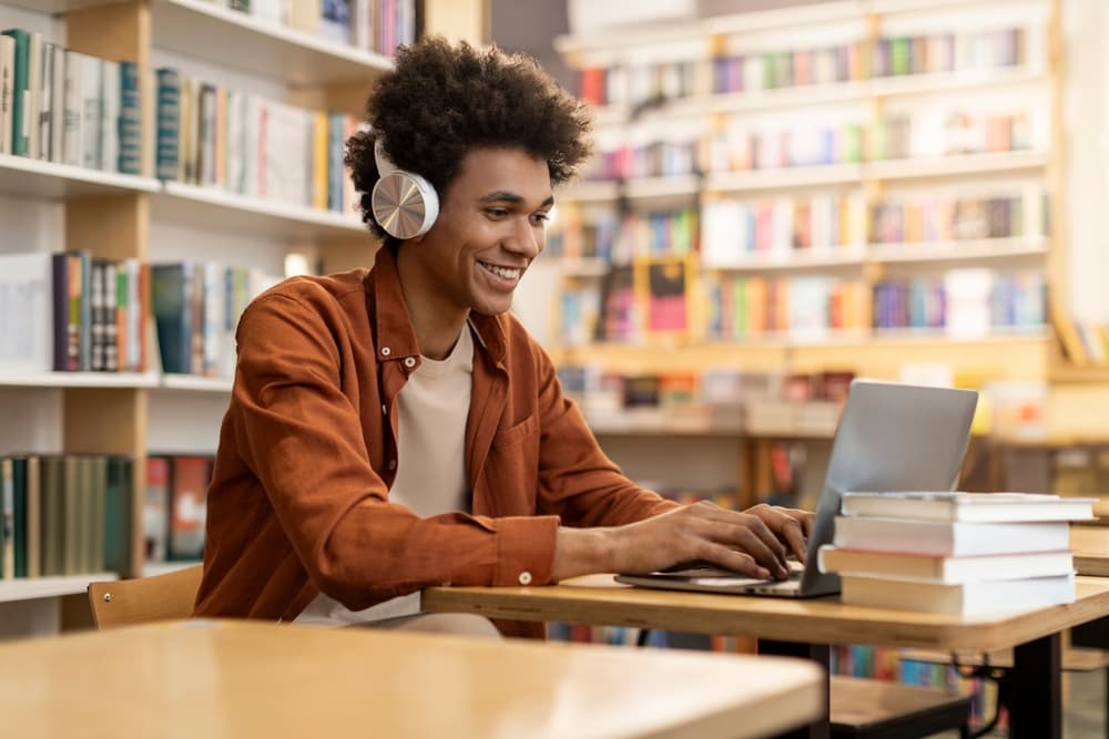 A smiling student listens to an online lecture on his laptop while working in a library.