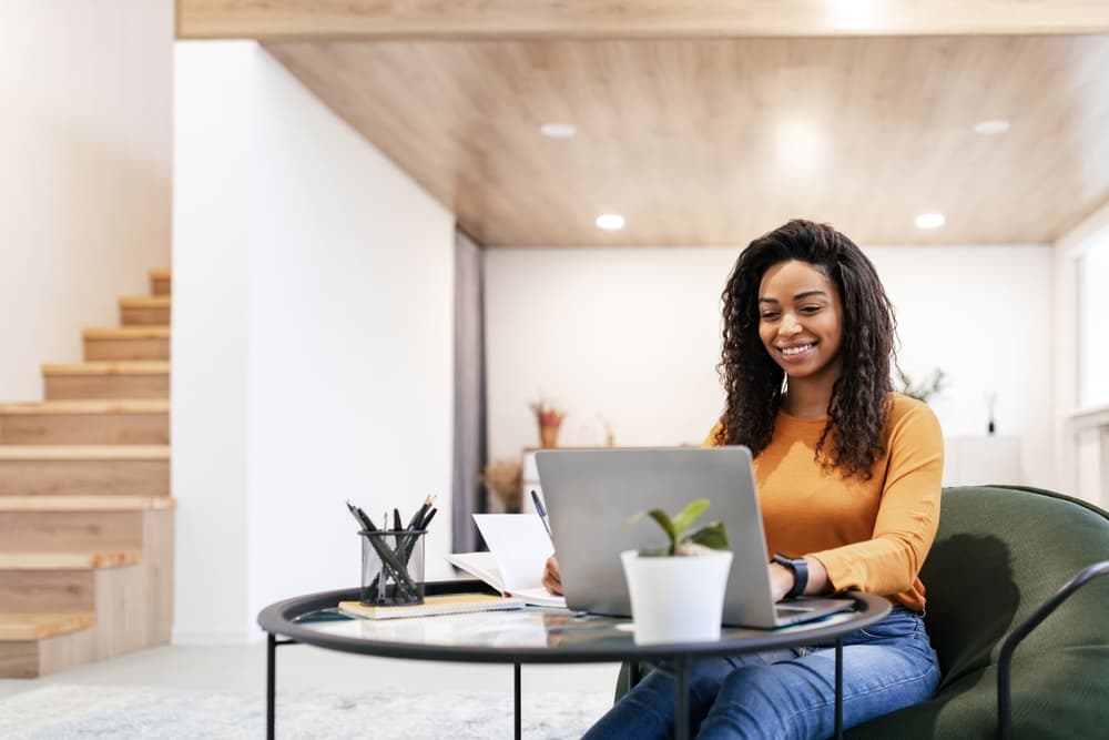 A smiling African American woman sits at a table in her home while working on her laptop.