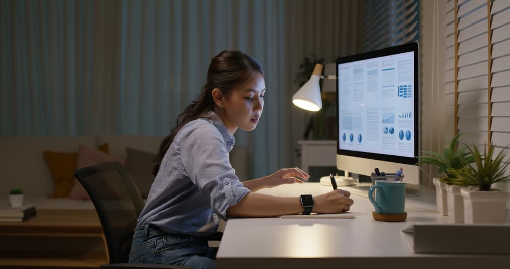 A female marketing research analyst reviews data on her computer while making notes in a notebook.