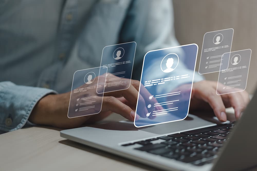 An HR manager’s hands are working on his laptop as he reviews potential candidates. The image superimposes the online CV cards of potential recruits.