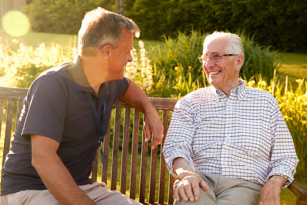 An assisted living facility manager chats with a smiling senior resident as they sit outside on a sunny day.
