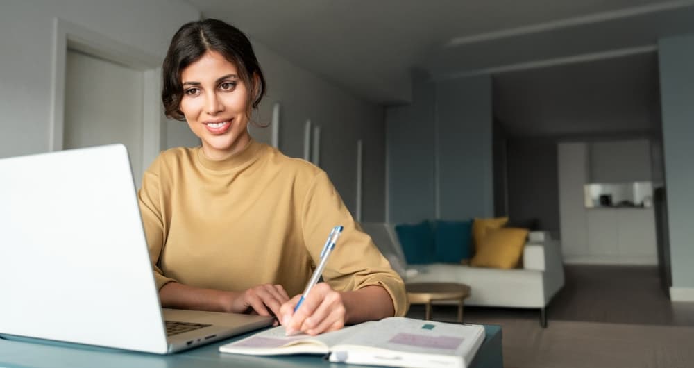 A smiling female adult student sits at her home desk, taking notes while reviewing an online lecture.