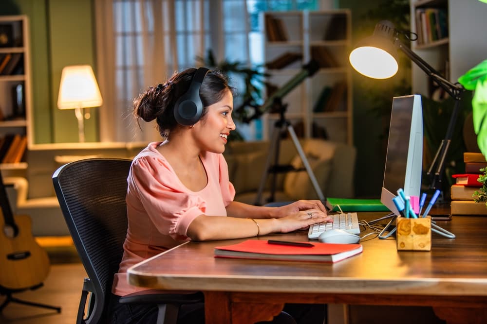 A smiling woman sits at her desk at home, wearing headphones and listening to an online lecture on her computer.