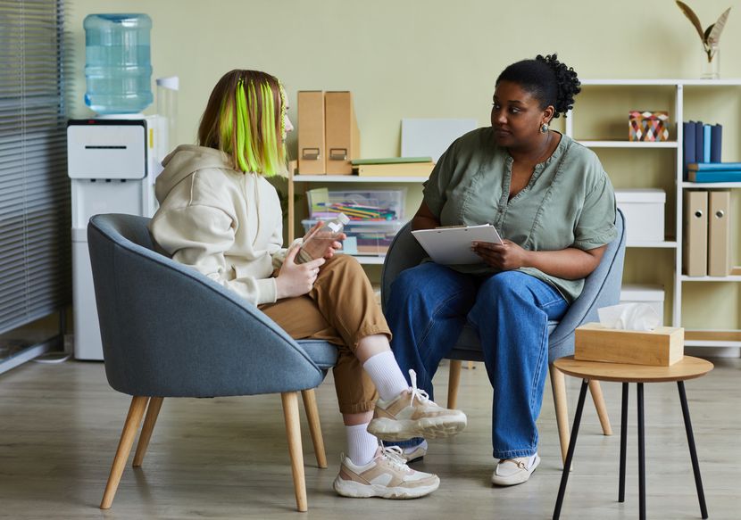 African female social worker talking to difficult teenage girl while they sitting on armchairs at office