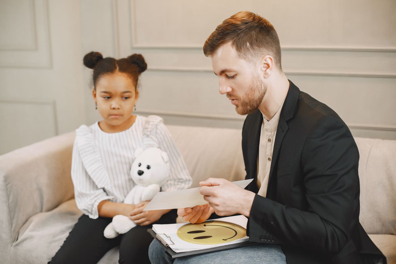 A family therapist reviews a drawing during a session with a young girl who sits beside him on a couch holding a white teddy bear.