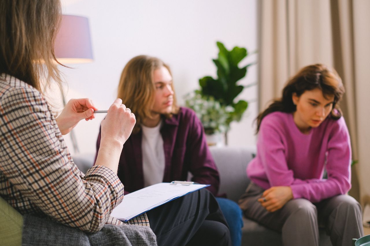 A marriage and family therapist holding a clipboard speaks with a young couple seated apart on a couch, the woman leaning forward with a troubled expression.