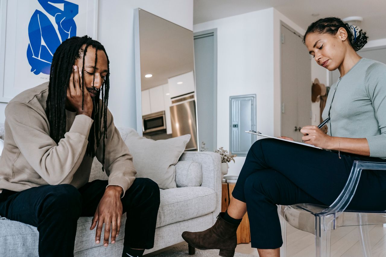 A man sits on a couch with his head resting in his hand while a counselor takes notes on a clipboard during a therapy session.