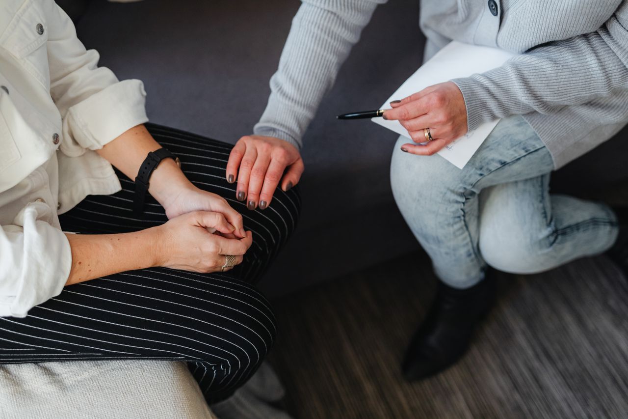 A marriage and family therapist holding a pen and notes reaches out and rests a comforting hand on a client’s knee during a therapy session.