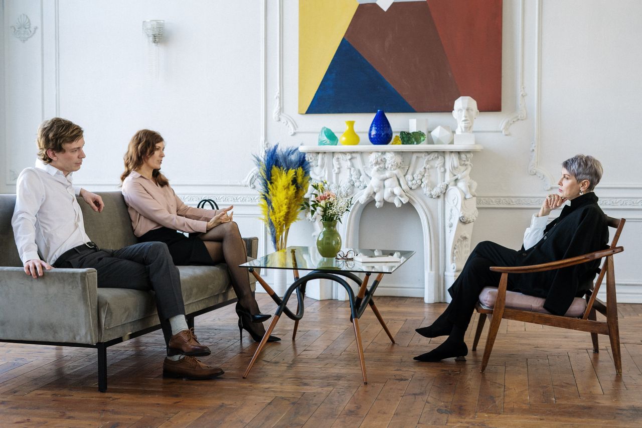 A marriage and family therapist sits in an armchair listening thoughtfully as a couple on a sofa across from her talks, the woman gesturing as she speaks.